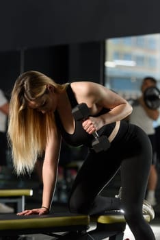 Athlete woman in sportswear training back with dumbbell in one hand leaning on the bench at fitness gym