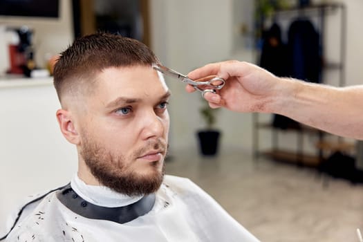 hairdresser does haircut for caucasian bearded man using comb and grooming scissors in barber shop.