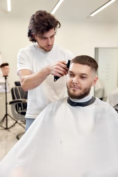 Barber trim hair with clipper on handsome bearded man in barber shop.