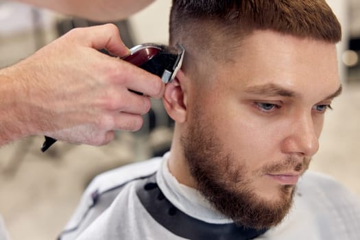 Barber trim hair with clipper on handsome bearded man in barber shop.