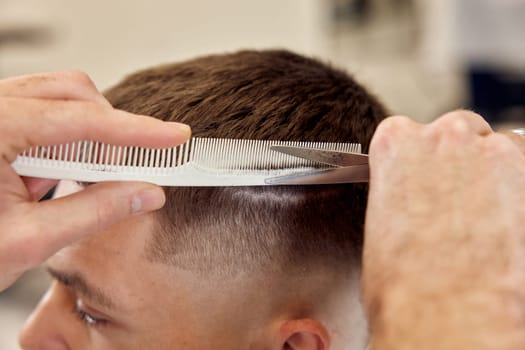 Professional hairdresser does haircut for caucasian bearded man using comb and scissors at barber shop. close-up