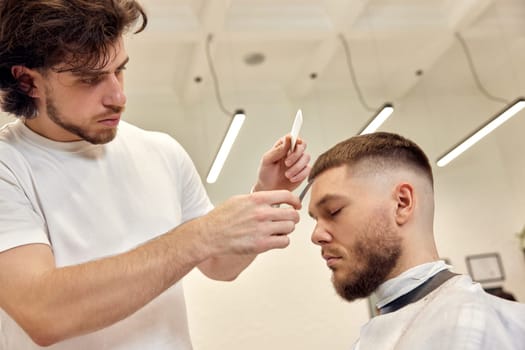 Professional hairdresser does haircut for caucasian bearded man using comb and scissors at barber shop. close-up