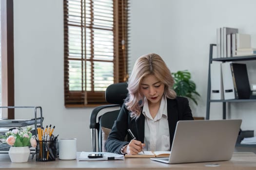 Portrait of smiling business woman in sitting at desk, using laptop and writing in notebook tutorial.