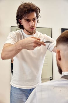 Professional hairdresser does haircut for caucasian bearded man using comb and scissors at barber shop.
