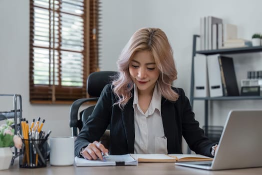 Portrait of smiling business woman in sitting at desk, using laptop and writing in notebook tutorial.