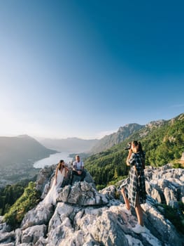 Girl-photographer photographs bride and groom sitting on a rock in the mountains above the Kotor Bay. High quality photo
