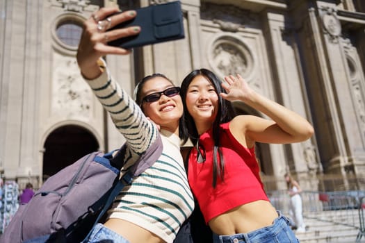 Cheerful young Asian female tourists in casual outfits smiling and taking self portrait on smartphone while standing against Cathedral of Granada in Spain during holidays