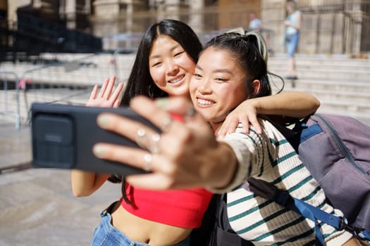 Happy young Asian female travelers, in casual clothes with backpacks smiling and hugging while waving hands and taking selfie on smartphone during summer vacation in Spain