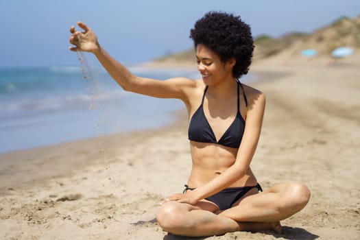 Full body of positive African American female traveler in bikini pouring sand while sitting on coast near sea in tropical resort in sunny weather