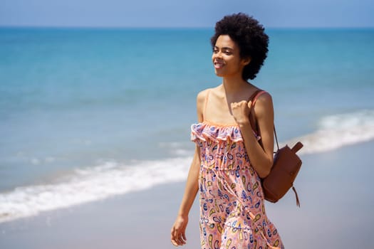 Black woman in colorful dress smiling while exploring sandy beach near rippling ocean and looking away