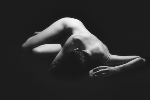 Full body of anonymous fit naked woman with short dark hair lying on floor against black background in studio. Black and white photograph