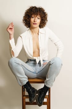 Full length of flirty female model with curly hair, in stylish wear resting on wooden stool playing with curl and looking at camera against white background