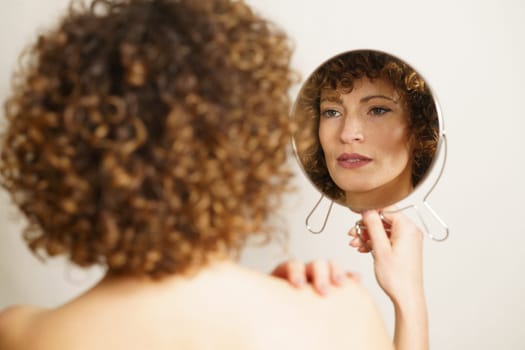Beautiful young woman, with curly hair and makeup standing with back against camera while holding mirror in hand and looking away in reflection of self near gray background