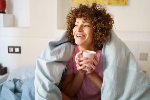 Satisfied female with cup of hot drink looking away while resting on bed in morning at home