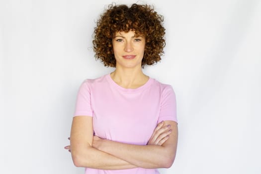 Curly haired woman in casual clothes standing with crossed hands and looking at camera while smiling in light studio