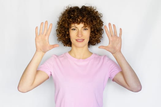 Attractive curly haired girl in casual outfit raising hands and showing number ten while looking at camera on white background