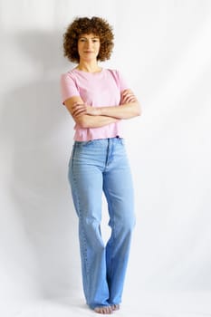 Full body of curly haired woman in casual clothes leaning on white wall while standing with hands crossed looking at camera and smiling