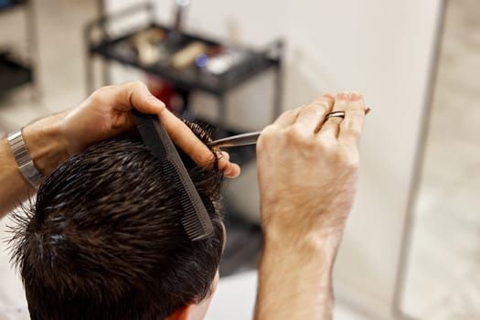 close-up, professional hairstylist does haircut for caucasian client man using comb and scissors at barber shop.