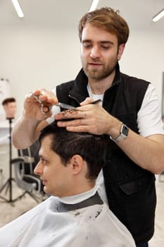 Professional hairdresser does haircut for caucasian client man using comb and scissors at barber shop.