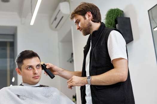 Barber cut hair with electric shearer machine on man in barber shop.