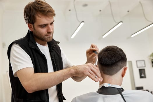 Professional hairdresser does haircut for caucasian bearded man using comb and scissors at barber shop.