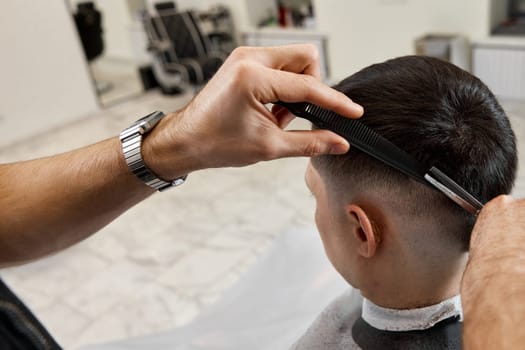 Professional hairdresser does haircut for caucasian bearded man using comb and scissors at barber shop.