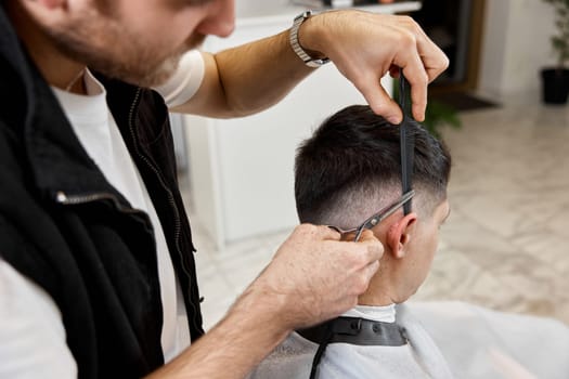 Professional hairdresser does haircut for caucasian bearded man using comb and scissors at barber shop.