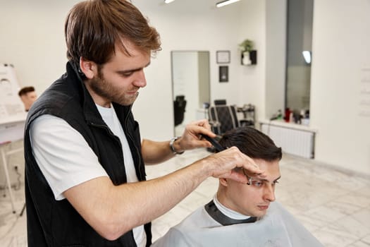 Professional hairdresser does haircut for caucasian bearded man using comb and scissors at barber shop.