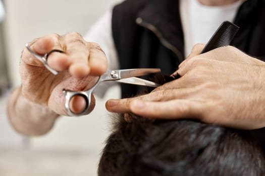 Professional hairdresser does haircut for caucasian bearded man using comb and scissors at barber shop.