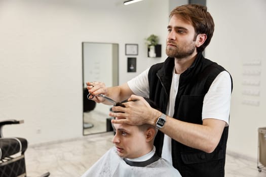 Professional hairdresser does haircut for caucasian bearded man using comb and scissors at barber shop.