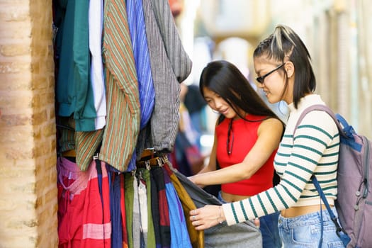 Side view of Asian young female friends in casual clothes buying clothes while shopping on street market in city during vacation