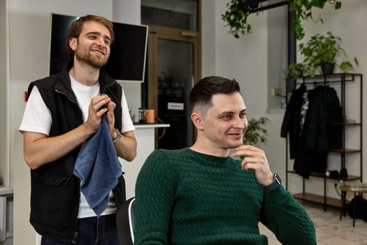 Barber talking to satisfied caucasian client man while sitting in chair after haircut at barbershop