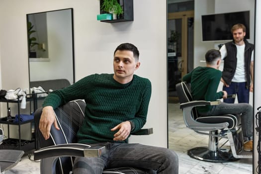 satisfied caucasian client man talking with barber while sitting in chair after haircut at barbershop