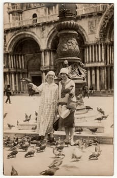 VENEZIA, ITALY - CIRCA 1926: Vintage photo shows women feed the pigeons on the square in Venezia. Women wear cloche hats. Vacation theme.