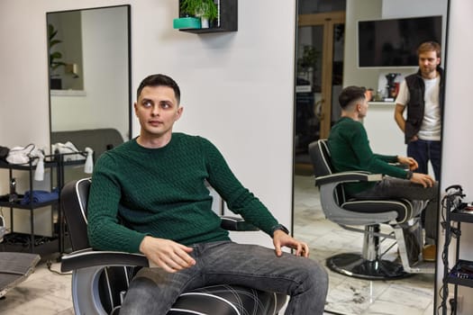 satisfied caucasian client man talking with barber while sitting in chair after haircut at barbershop