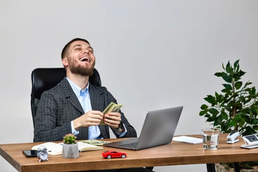 young bearded businessman working on laptop and counting dollars while sitting on chair at desk.