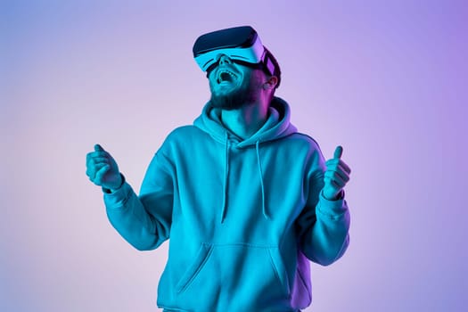 man in sweatshirt using virtual reality glasses and playing video games on blue and pink neon background.