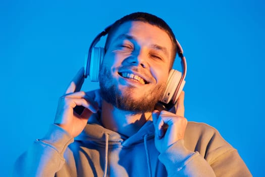 handsome bearded man with headphones in sweatshirt enjoying favorite music on blue neon background. Neon lighting