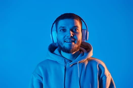 handsome bearded man with headphones in sweatshirt enjoying favorite music on blue neon background. Neon lighting