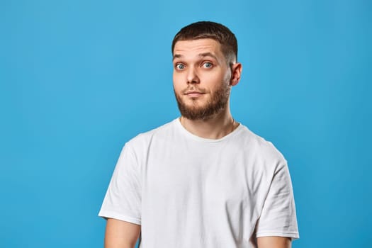 puzzled young bearded man in casual white t-shirt looking at the camera on blue gray background.