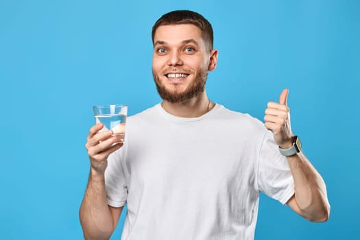 handsome bearded man in white t-shirt with drinking glass of water showing thumbs up gesture on blue background