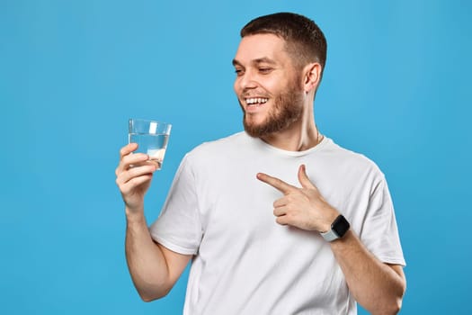 positive handsome man in white t-shirt showing drinking glass of water on blue background