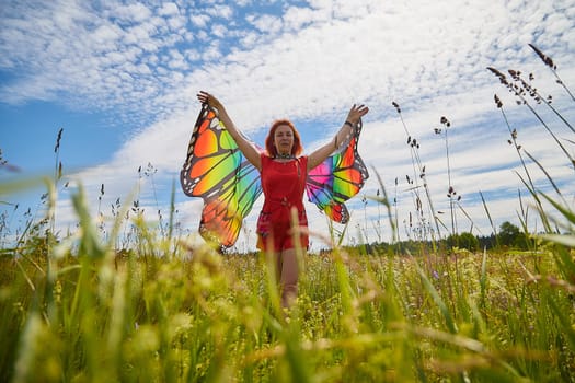 Adult girl with red hair and butterfly wings having fun and joy in meadow or field with grass, flowers on sunny summer day