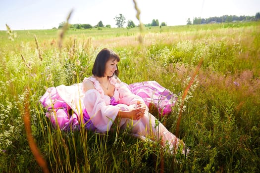 Beautiful girl in lush pink ball gown in green field during blooming of flowers and blue sky on background. Model posing on nature landscape as princess from fary tale