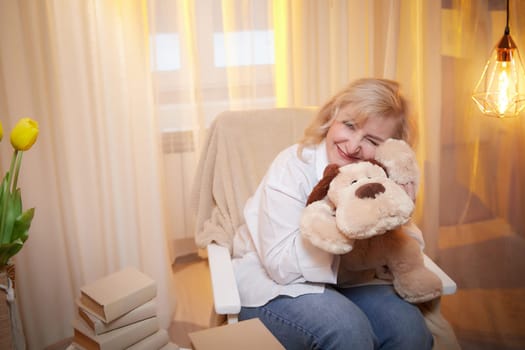 Adult mature woman of 40-60 years old with funny dog toy in a casual dress with white shirt and jeans. Room with calm cozy evening atmosphere with transparent curtains and soft warm light of lamp