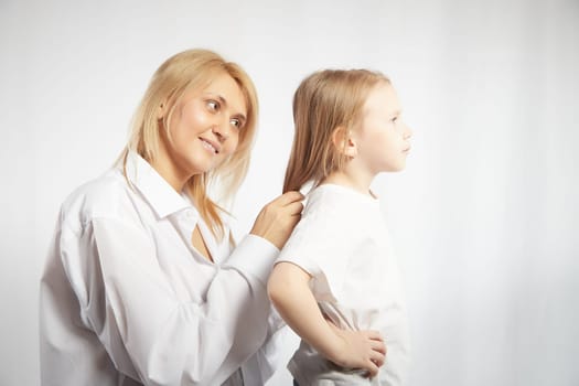 Close portrait of blonde mother and daughter where mom braids hair and makes ponytail and hairstyle for her girl on white background in studio. The concept of love, friendship, caring in family