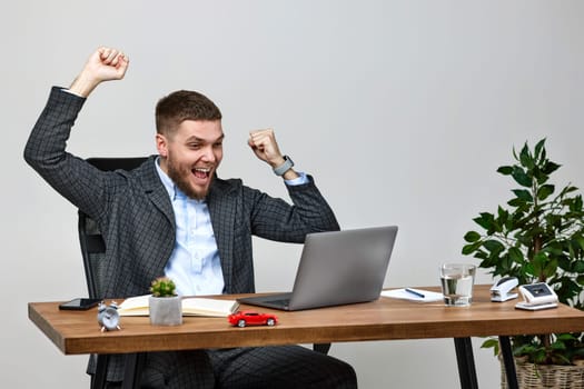 Satisfied young businessman gesturing emotionally and celebrating victory while sitting on chair at desk, using laptop pc computer