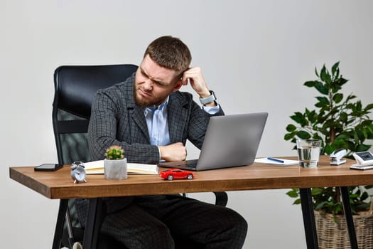 bearded man sitting on chair at desk and suffering from headache at workplace, using laptop