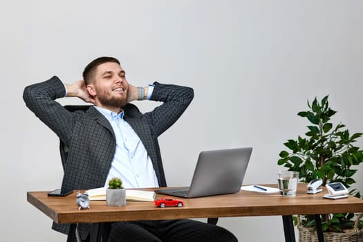 satisfied handsome bearded man holding hands behind head, sitting on chair at table and resting, using laptop