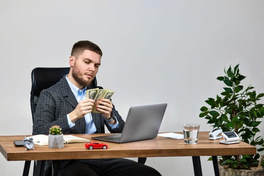 young bearded businessman working on laptop and holding money while sitting on chair at desk.